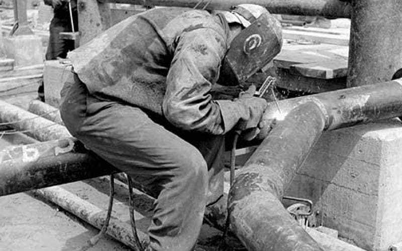 Male welder wearing welding mask joins Y-shaped steam pipe in the Polymer Corporation Limited plant, Sarnia, Ontario (3…